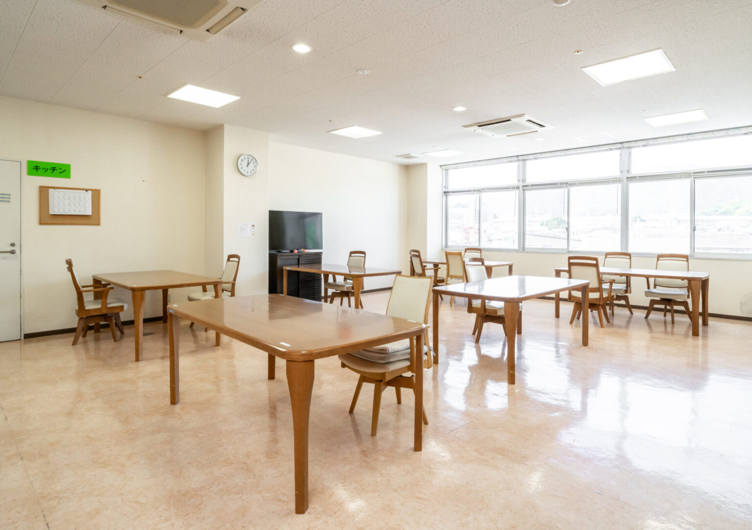 明るい共用ラウンジ。複数のテーブルと椅子、テレビ、大窓が備わった食事・交流スペース。