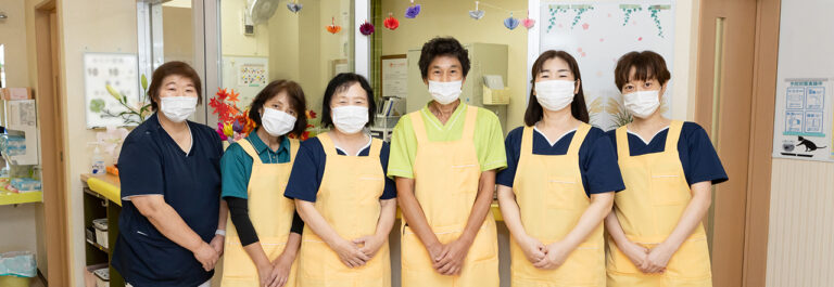 黄色いエプロンを着けたスタッフ6名が揃って笑顔で撮影に応じている職員写真